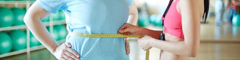 weight management concept - trainer measuring client's waist line with measuring tape